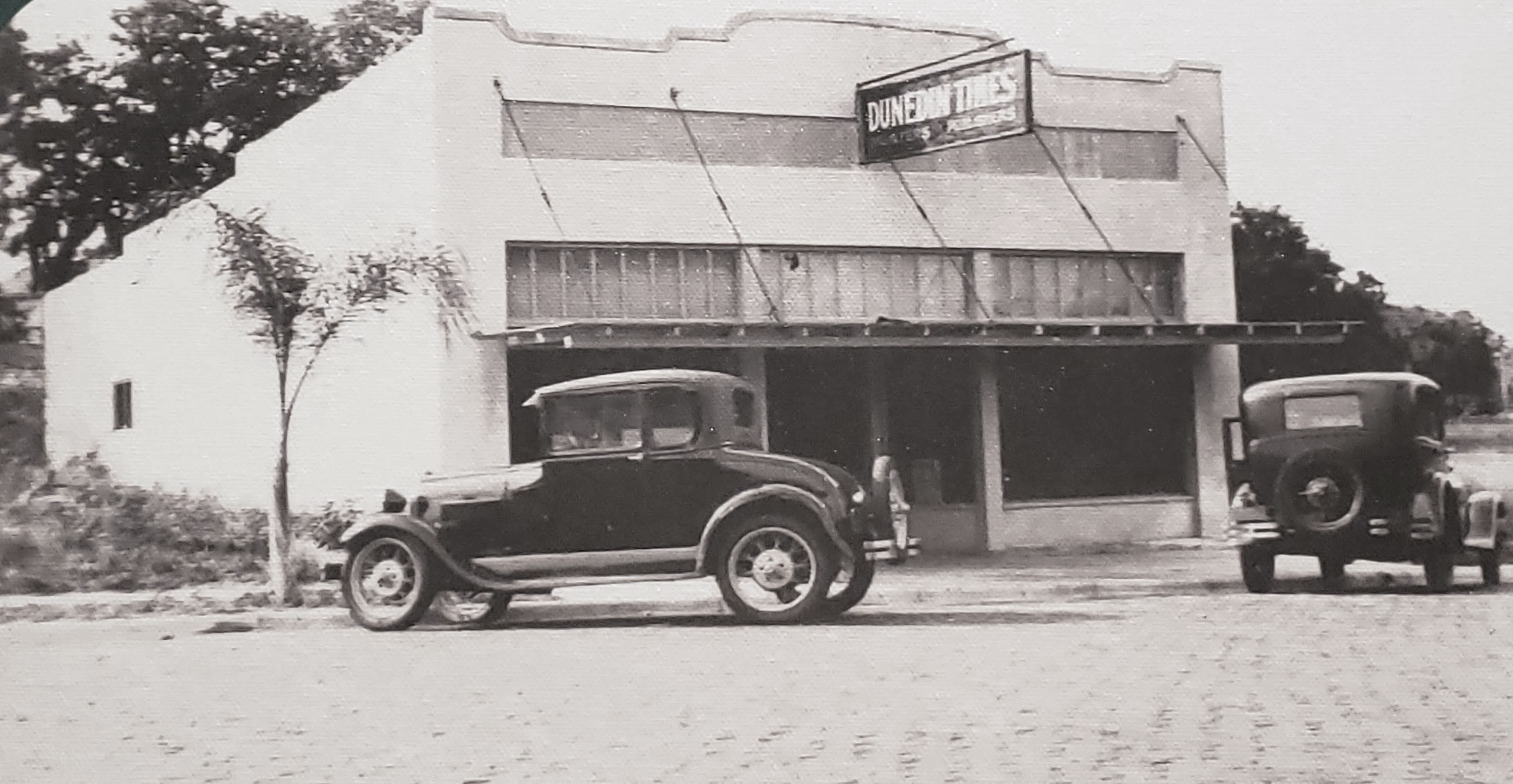 Historical black and white photo of the single-story Dunedin Times Newspaper building with a wooden hanging sign above the entrance and two vintage Ford Model T cars parked in front.