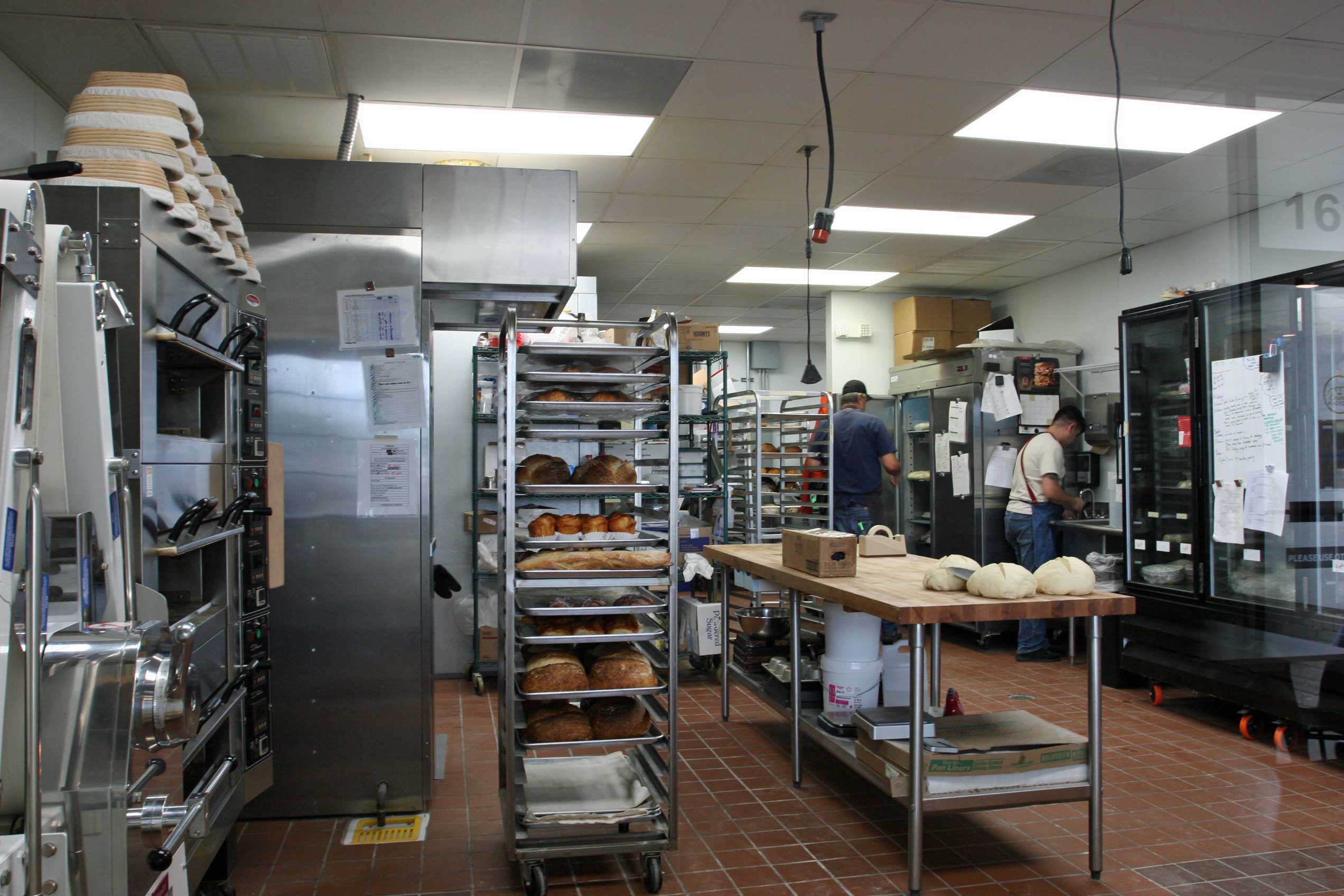 Bakers working in the background behind a wooden butcher block work table with proofing dough, surrounded by kitchen tools, a bread oven, and racks of freshly baked bread.
