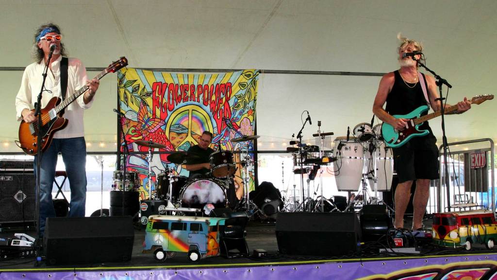 The band St. Pete Sons performing live on the festival stage. In the center, the drummer plays his kit, flanked on either side by two bass players caught mid-performance in energetic, singing action poses.