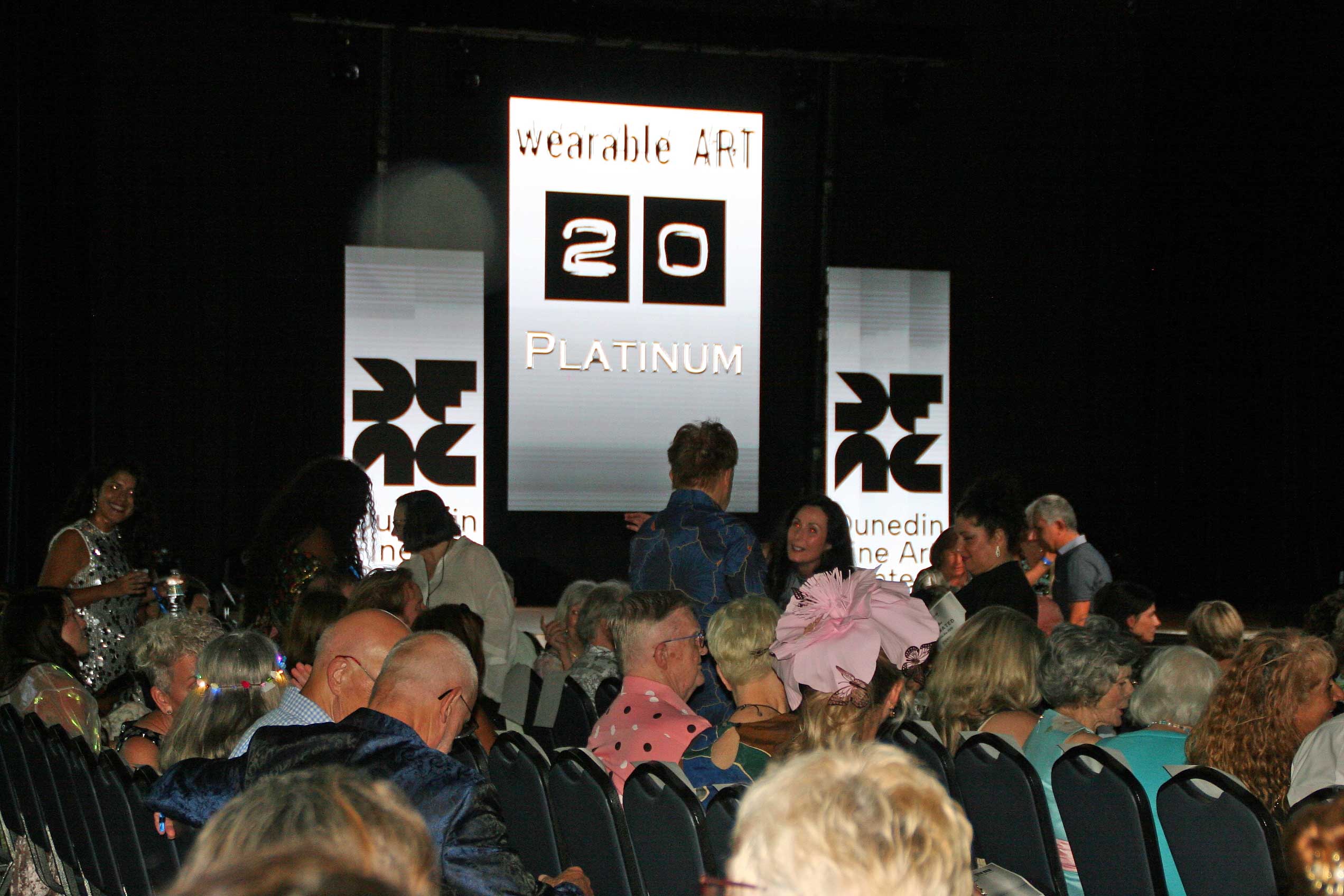 A wide shot of the wearable ART 20 Platinum fashion runway at the Dunedin Fine Art Center, illuminated by vibrant stage lighting with guests seated along the runway.