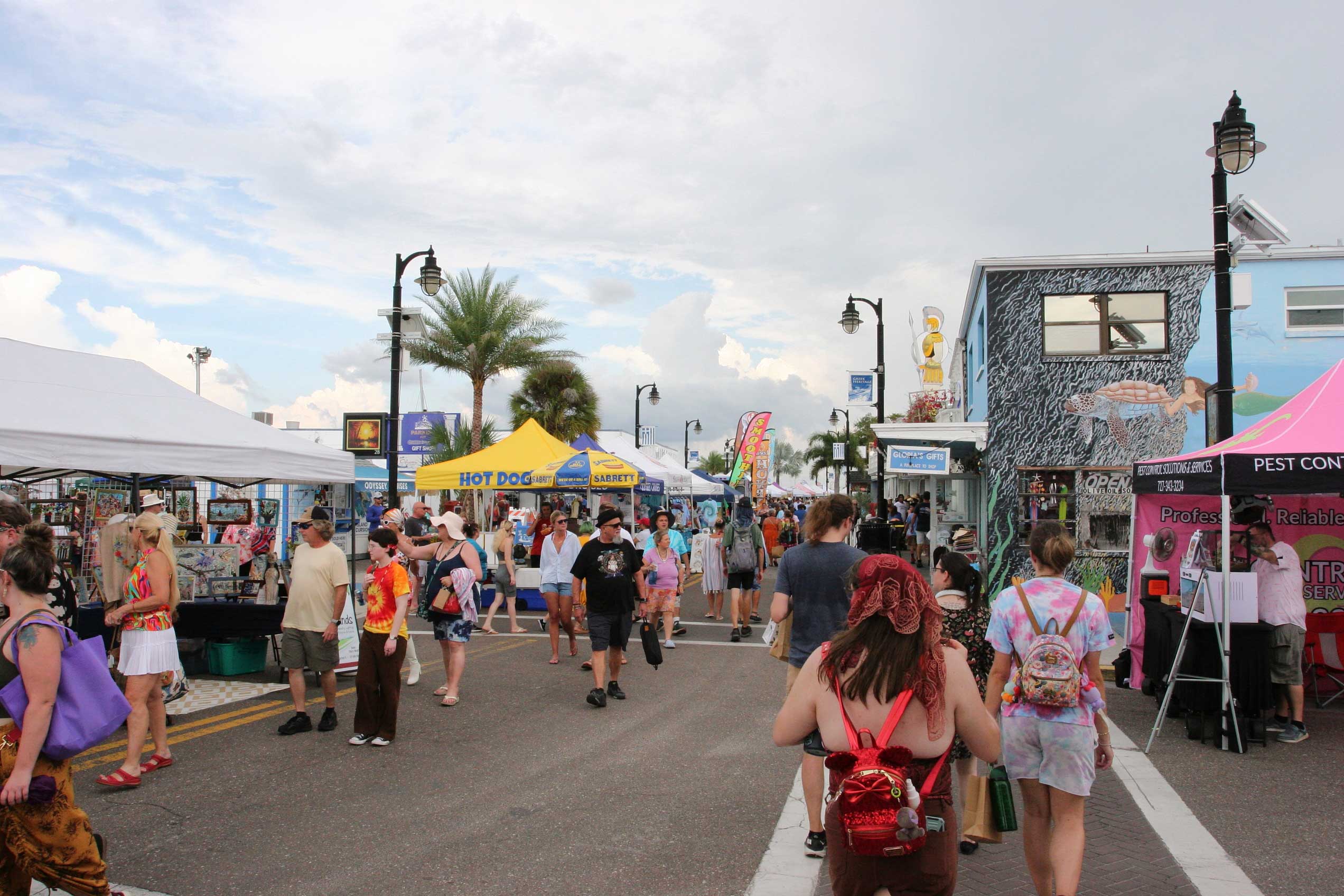 A lively street-level view along Dodecanese Boulevard during the festival, showing several vendors under pop-up tents selling products on the street. Crowds of shoppers walk from tent to tent, while other pedestrians stroll along the sidewalk passing local storefronts.