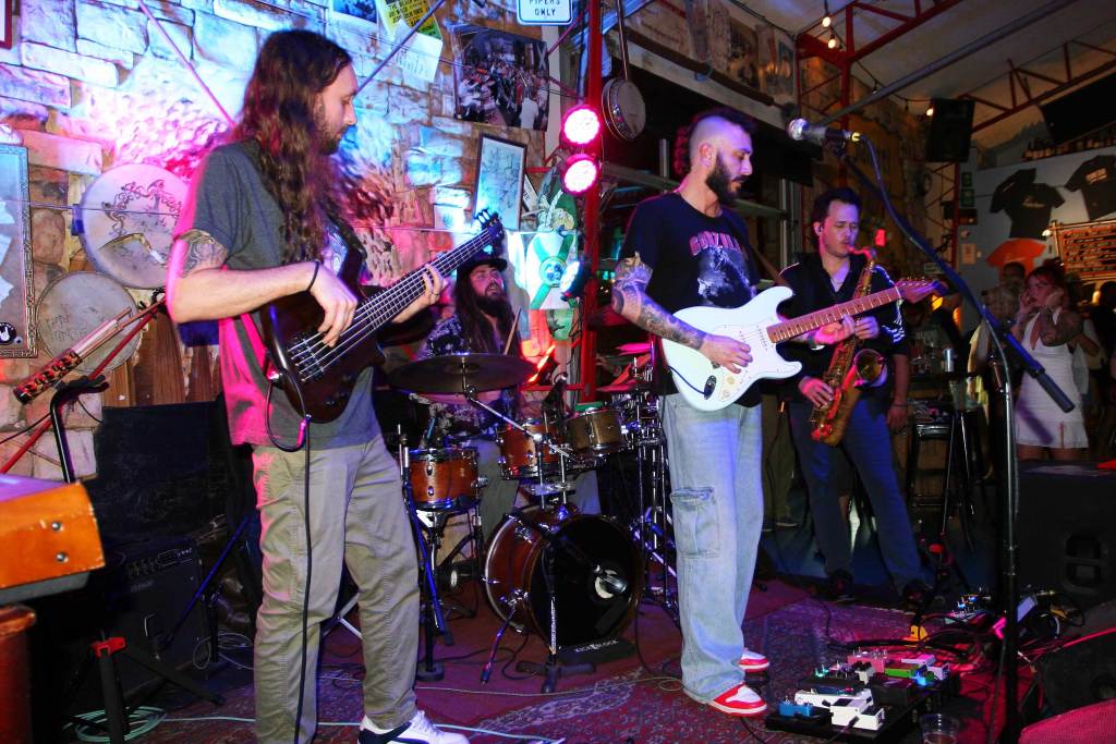 Instrumental band Sauce Pocket performing live on stage with two electric guitarists, a saxophonist, and a drummer in the background.