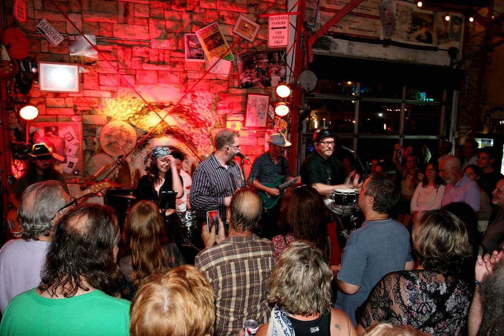 Talking Heads tribute band Road To Nowhere performing live on a packed stage. Three lead vocalists, including one woman and one man playing bongo drums, stand in the foreground, with two bass players and a drummer positioned in the background.
